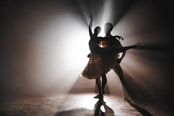 Graziosa ballerina e il suo partner maschile ballano elementi di balletto classico o moderno al buio con retroilluminazione. Coppia in fumo su sfondo nero. Concetto artistico . — Foto Stock