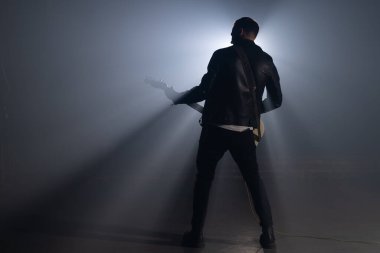 Deri ceketli, dumanlı stüdyoda rock gitaristi ya da sahne ustaca elektro gitar çalıyor. Sahne ışıkları altında müzisyen görüntüsü.