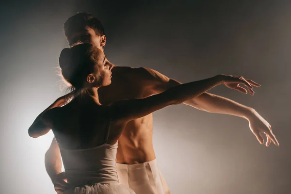 Bailarina graciosa e seu parceiro masculino dançando elementos de balé clássico ou moderno no escuro com luz de fundo de holofotes. Casal em fumaça no fundo preto. Conceito de arte . — Fotografia de Stock