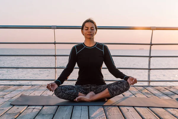 Ragazza concentrata in nero pratica nella meditazione yoga sul lungomare di legno. Donna in posa di loto. Allungamento, pratica, stile di vita sano. Copia spazio — Foto Stock