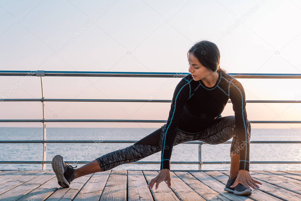 Young athletic girl performs squats on wooden embankment by the sea in early morning. Healthy lifestyle, coaching, training concept.