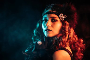 Flappers tarzı giyinmiş eski moda bir kadın. Elinde sigarayla karanlık arka planda, neon ışıklı bir ağızlık ile poz veriyor. Kükreyen yirmiler, retro, parti, moda konsepti