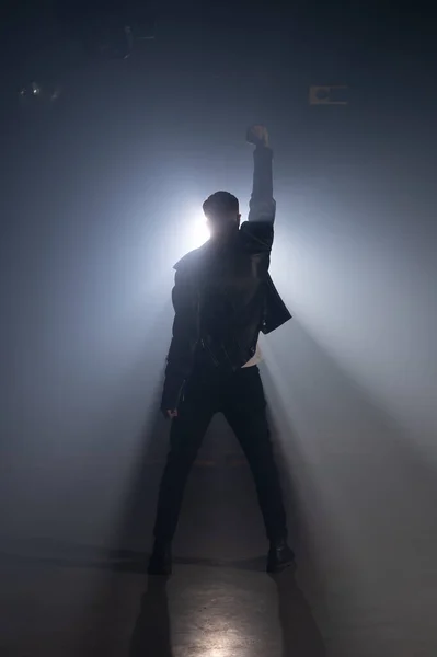 Young man in leather jacket on smoky stage. View of guy raises hand up as gesture and symbol of success, freedom and victory. — Stock Photo, Image