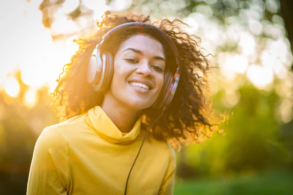 Close-up of african american woman listening to music with headphones outdoors. Young girl with curly hairstyle in yellow enjoys music in park.