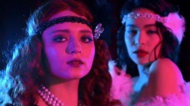 Muhteşem Gatsby tarzı giyinmiş, pembe neon ışıkta koyu arka planda poz veren genç kadınların portresi. Kükreyen yirmiler, retro, parti, moda konsepti