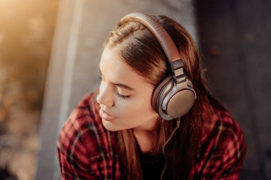 Genç kız kulaklıkla müzik dinliyor. Kırmızı ekose gömlekli kız gülümsüyor, ritimle dans ediyor. Öğrenci hayatı, özgürlük, modern gençlik kavramı.