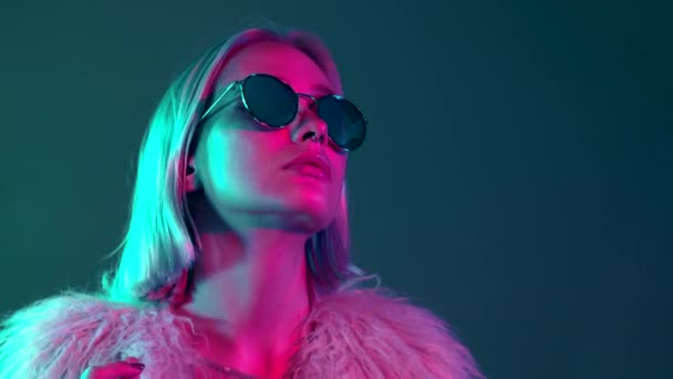 Glamorous hipster teenager in sunglasses and furry coat dancing to music with sunglasses. Portrait of millennial pretty girl with short hairstyle with neon light. Dyed blue and pink hair. — Stock Video