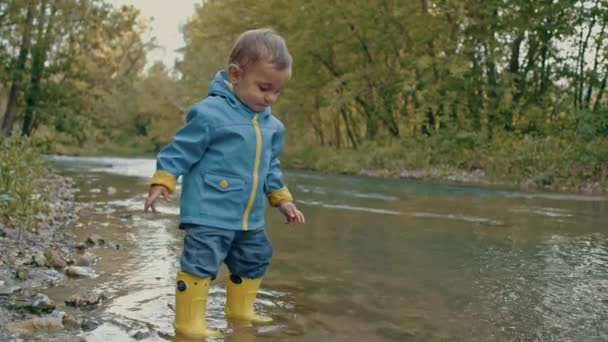 Aranyos baba esőkabátban és gumicsizmában szórakozik a folyónál, kavicsokat dobál és nevet. Vicces fiú tanul és felfedezi a természet világát. Család, gyerekek, szerelem koncepció. — Stock videók