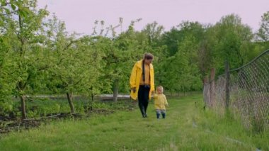 Sarı yağmurluk giyen anne ve yakışıklı bebek yağmurdan sonra yeşil bahçede yürüyorlar. Çocuk annelerinin elini tutuyor. Aşk, ilgi, bağlılık, aile, çocuk konsepti.