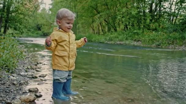 Aranyos baba esőkabátban és gumicsizmában szórakozik a folyónál, kavicsokat dobál és nevet. Vicces fiú tanul és felfedezi a természet világát. Család, gyerekek, szerelem koncepció. — Stock videók