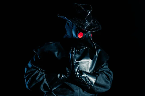 Man in plague doctor costume with crow-like mask praying with hands isolated on black background. Creepy mask, historical costume concept. Epidemic