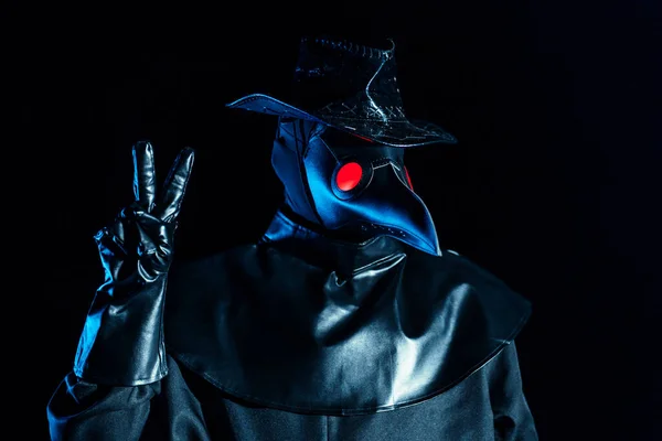 Man in plague doctor costume with crow-like mask showing peace gesture isolated on black background. Creepy mask, historical costume concept. Epidemic
