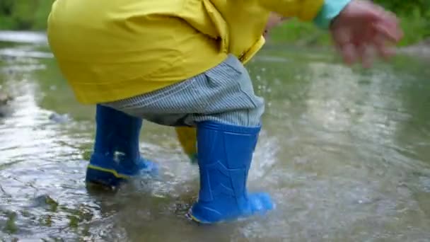 Bebé en impermeable y botas de goma se divierte por el río, tocando el agua con las manos. Chico divertido aprende y explora el mundo natural. Familia, niños, concepto de amor . — Vídeos de Stock