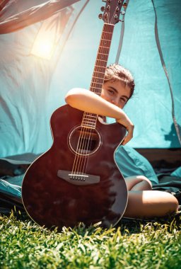 Genç kız taze yeşil çimlerde gitarla oturuyor, yetenekli çocuk müzik aleti çalıyor, yaz kampında eğleniyor.