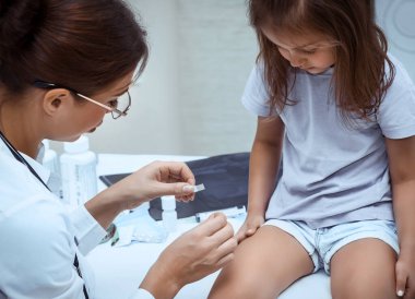 Pediatri randevusundaki tatlı küçük kız, beyaz doktor önlüklü hoş bayan doktor, kızın dizine yara bandı yapıştırıyor, hastanedeki çocuk.