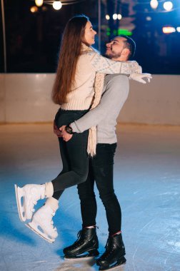 Couple on the city rink in a winter evening. Guy helping nice girl to skate on the ice in the dark night and twinkles lighting above them