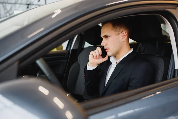 Young businessman sits in luxury car and talks on phone. He looks straight forward. Guy drives car. He holds one hand on steering wheel. It is sunny outside.
