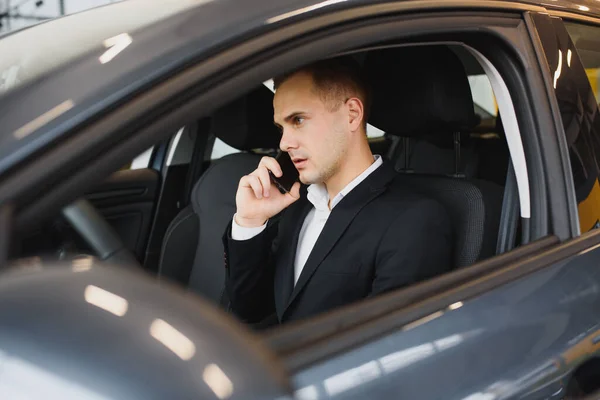 Young businessman sits in luxury car and talks on phone. He looks straight forward. Guy drives car. He holds one hand on steering wheel. It is sunny outside.