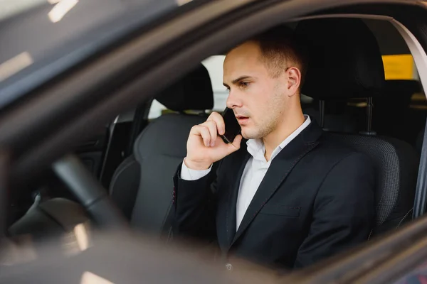 Young businessman sits in luxury car and talks on phone. He looks straight forward. Guy drives car. He holds one hand on steering wheel. It is sunny outside.