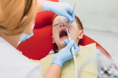 Pediatrik dişçi, diş kliniğindeki dişçi koltuğunda küçük bir çocuğun dişlerini inceliyor.