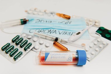 CAVID-2019 Coronavirus için pozitif bir test tüpü. Tıbbi aletler beyaz bir masada. Termometre, fonendoskop, haplar, cerrahi maskeler.