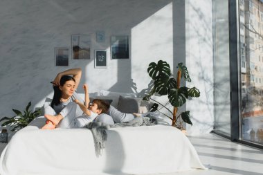 Genç anne ve 4 yaşındaki oğlu pijama giyip hafta sonu yatakta oynuyorlar. Tembel sabah, sıcak ve rahat bir ortam..