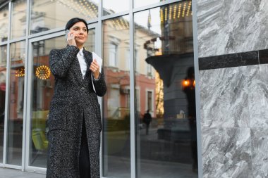 Şehirdeki çekici genç kadın. İş kadını, modern iş merkezinin önünde akıllı bir telefonla konuşurken gülümsüyor.