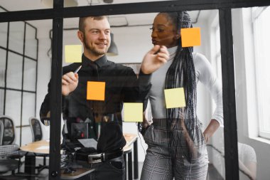 Gülümseyen takım liderinin ofisteki Kanban çevik cam pencere tahtasının renkli yapışkan notlarıyla yaptığı görevi izleyerek mutlu melez yarış arkadaşları. Çeşitli takım arkadaşları çalışma sürecinden memnun.