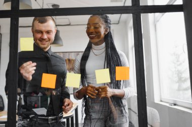 Gülümseyen takım liderinin ofisteki Kanban çevik cam pencere tahtasının renkli yapışkan notlarıyla yaptığı görevi izleyerek mutlu melez yarış arkadaşları. Çeşitli takım arkadaşları çalışma sürecinden memnun.