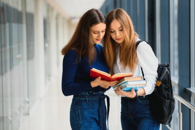Koridorda kitap ve çantalarla dikilen iki genç kız öğrenci birbirleriyle konuşuyor..