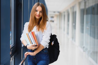 Siyah sırt çantalı neşeli esmer kız öğrenci modern binada kitapları tutuyor. Üniversite koridorunda elinde kitaplarla duran kız öğrenci.