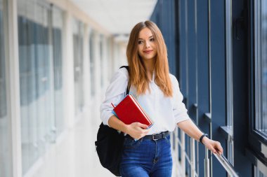 Siyah sırt çantalı neşeli esmer kız öğrenci modern binada kitapları tutuyor. Üniversite koridorunda elinde kitaplarla duran kız öğrenci.