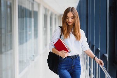 Siyah sırt çantalı neşeli esmer kız öğrenci modern binada kitapları tutuyor. Üniversite koridorunda elinde kitaplarla duran kız öğrenci.
