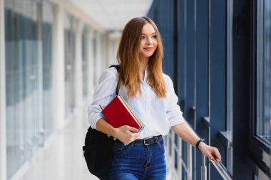 Siyah sırt çantalı neşeli esmer kız öğrenci modern binada kitapları tutuyor. Üniversite koridorunda elinde kitaplarla duran kız öğrenci.