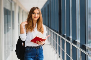 Siyah sırt çantalı neşeli esmer kız öğrenci modern binada kitapları tutuyor. Üniversite koridorunda elinde kitaplarla duran kız öğrenci.