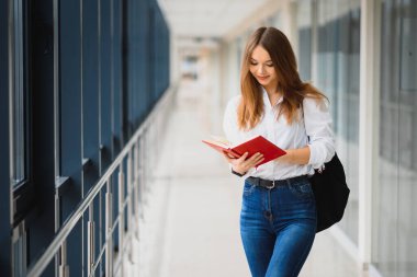 Siyah sırt çantalı neşeli esmer kız öğrenci modern binada kitapları tutuyor. Üniversite koridorunda elinde kitaplarla duran kız öğrenci.