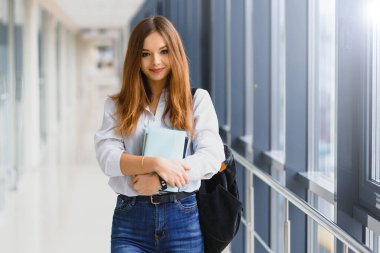 Siyah sırt çantalı neşeli esmer kız öğrenci modern binada kitapları tutuyor. Üniversite koridorunda elinde kitaplarla duran kız öğrenci.