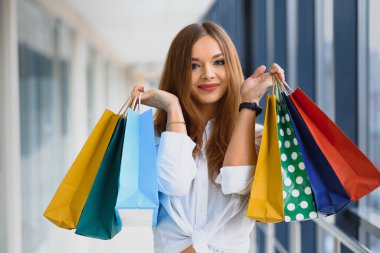 Alışveriş torbalı güzel kız alışveriş merkezinde alışveriş yaparken kameraya bakıyor ve gülümsüyor.
