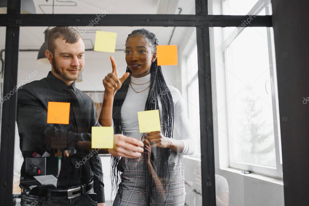 Diverse team of skilled students together solving task writing down information on papers glued on wall.Multicultural young people collaborating on business plan making notes of ideas on sticker