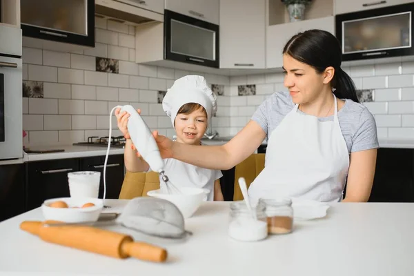 A young and beautiful mom is preparing food at home in the kitchen, along with her little son