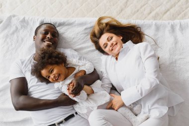 Evdeki rahat beyaz yatakta uzanan mutlu, çok ırklı bir ailenin portresi. Uluslararası anne ve baba, yatak odasındaki resim için poz veren melez bir kızla rahatlıyor.