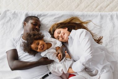 Evdeki rahat beyaz yatakta uzanan mutlu, çok ırklı bir ailenin portresi. Uluslararası anne ve baba, yatak odasındaki resim için poz veren melez bir kızla rahatlıyor.