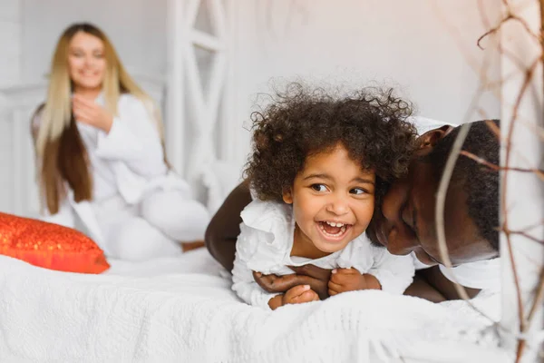 Portrait of happy multiracial young family lying on cozy white bed at home, smiling international mom and dad relaxing with little biracial girl child posing for picture in bedroom