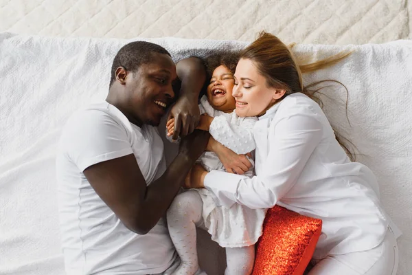 Portrait of happy multiracial young family lying on cozy white bed at home, smiling international mom and dad relaxing with little biracial girl child posing for picture in bedroom