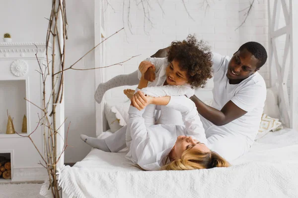 Portrait of happy multiracial young family lying on cozy white bed at home, smiling international mom and dad relaxing with little biracial girl child posing for picture in bedroom