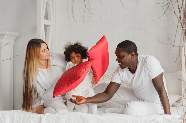 Portrait of happy multiracial young family lying on cozy white bed at home, smiling international mom and dad relaxing with little biracial girl child posing for picture in bedroom