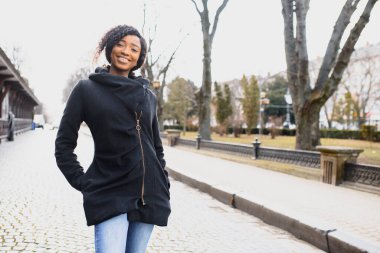 Afro saç stili ve burun halkası olan çekici, mutlu, koyu tenli bir model sabah yürüyüşü sırasında şehir arka planına karşı poz veriyor, beyaz dişlerini gösteren utangaç bir gülümsemeyle aşağıya bakıyor. Parlak güneş