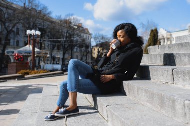 Neşeli, koyu tenli, kahve içmeyi seven, şehir parkında eğlenen, modaya uygun giyinmiş, Afro-Amerikan hippi kız güneşli bir günde dışarıda oturmuş içecek içiyor.