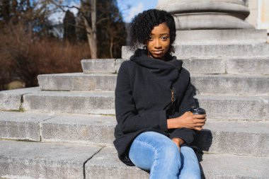 Neşeli, koyu tenli, kahve içmeyi seven, şehir parkında eğlenen, modaya uygun giyinmiş, Afro-Amerikan hippi kız güneşli bir günde dışarıda oturmuş içecek içiyor.