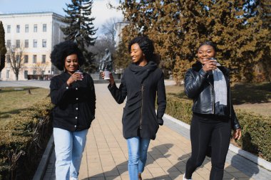 Üç güzel, koyu tenli, uzun saçlı kız bir şehirde duruyor, kahve içiyor ve telefonları kullanıyor.
